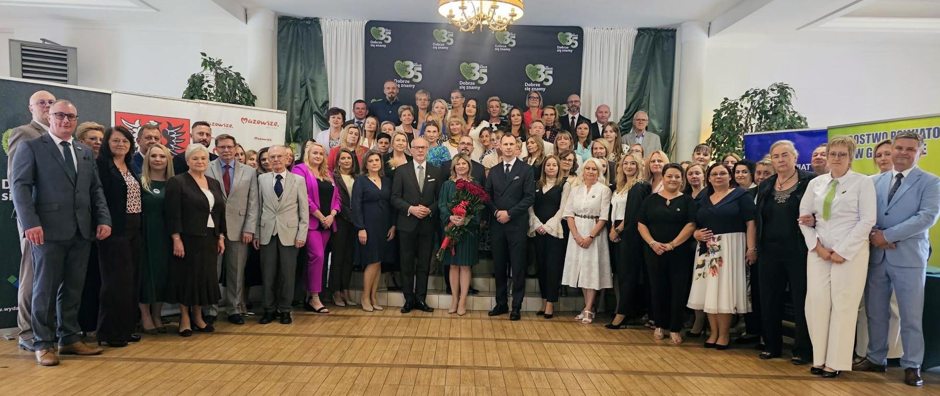 Kilkadziesiąt osób pozuje do zdjęcia stojąc na schodach w sali konferencyjnej udekorowanej ściankami i rollupami KRUS. Wszyscy uśmiechają się do zdjęcia.