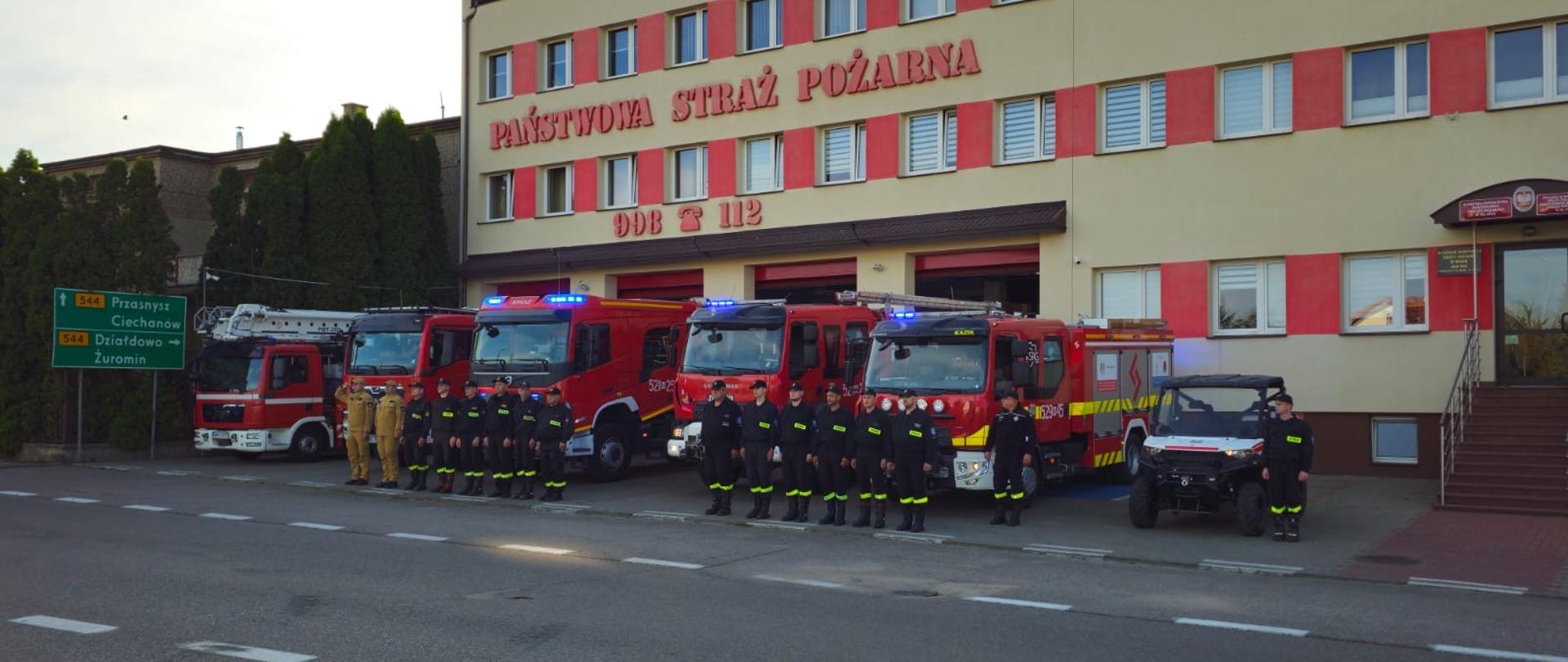 Przed budynkiem jednostki Państwowej Straży Pożarnej stoi szereg strażaków w umundurowaniu służbowym. W tle widoczne są cztery czerwone wozy strażackie z włączonymi niebieskimi światłami ostrzegawczymi, jeden pojazd specjalistyczny (prawdopodobnie drabina mechaniczna) oraz mały pojazd terenowy (quad typu UTV). Budynek straży pożarnej jest jasnożółty z czerwonymi akcentami wokół okien, a nad bramami garażowymi widnieje napis "PAŃSTWOWA STRAŻ POŻARNA", wraz z numerami alarmowymi 998 i 112. Po lewej stronie kadru znajdują się zielone tablice drogowe wskazujące kierunki do pobliskich miejscowości: Przasnysz, Ciechanów, Działdowo i Żuromin. Całość przedstawia formalne ustawienie służbowe przed jednostką, prawdopodobnie z okazji apelu, święta lub innego oficjalnego wydarzenia.