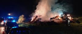 Zdjęcie przedstawia porę nocną z ciemnym tłem. Na pierwszym planie stoi kilka samochodów strażackich na drodze z włączonymi niebieskimi światłami błyskowymi, które oświetlają miejsce pożaru. Na drugim planie strażacy gaszący palącą się wysoką stertę słomy i koparka rozgarniająca słomę.