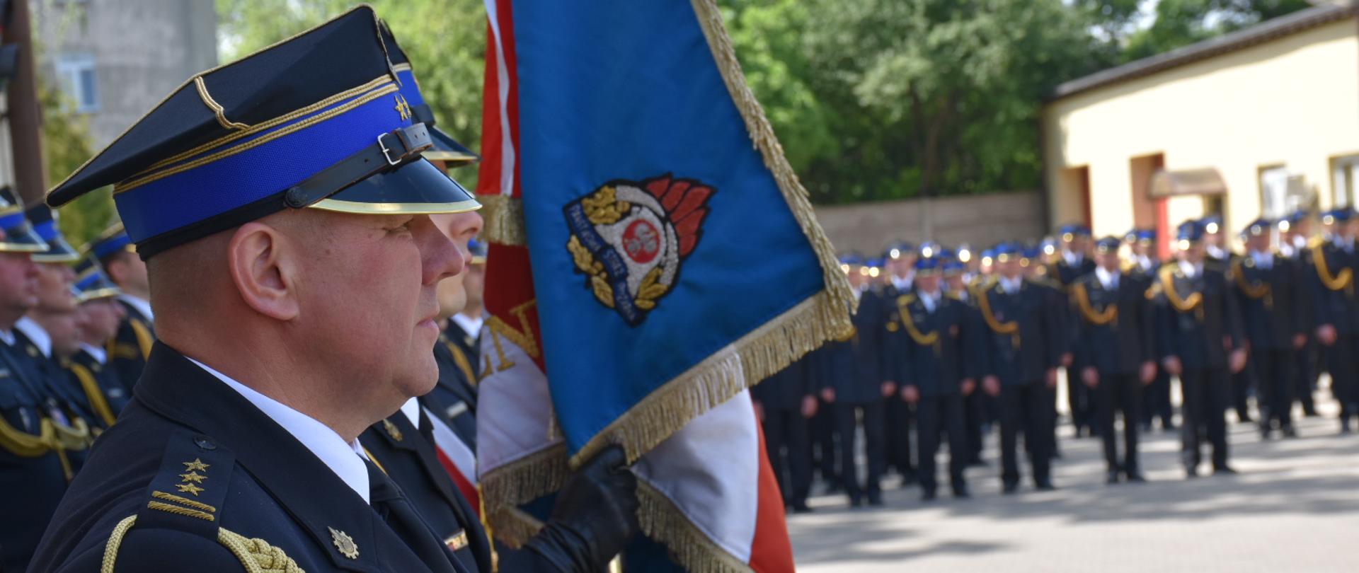 Zastępca komendanta miejskiego PSP stojący bokiem na czele pododdziału kadry kierowniczej KM PSP w Łodzi, w tle sztandar Komendy Miejskiej PSP w Łodzi 