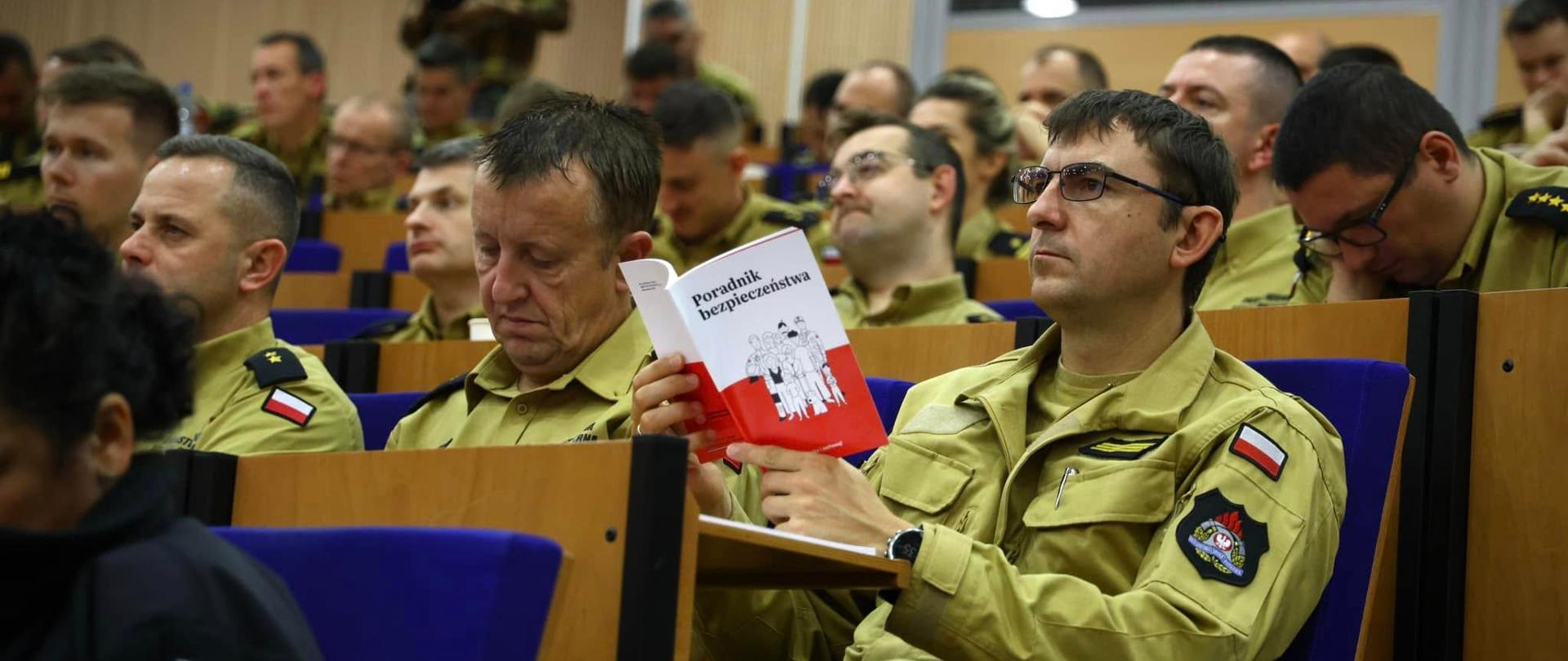 Na sali konferencyjnej, kaskadowo, siedzą strażacy w mundurach służbowych. Pierwszy z szeregu trzyma w ręku poradnik.