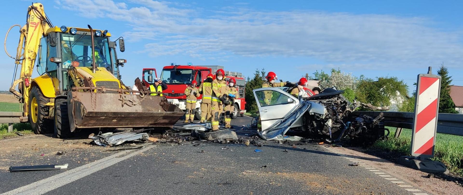 Kompletnie rozbity wrak samochodu osobowego przy barierce energochłonnej widziany boku. Na jezdni stoi również koparkoładowarka która zderzyła się z samochodem osobowym. W tle widoczny wóz strażacki i pracujący strażacy, ratownicy medyczni i policjanci. 