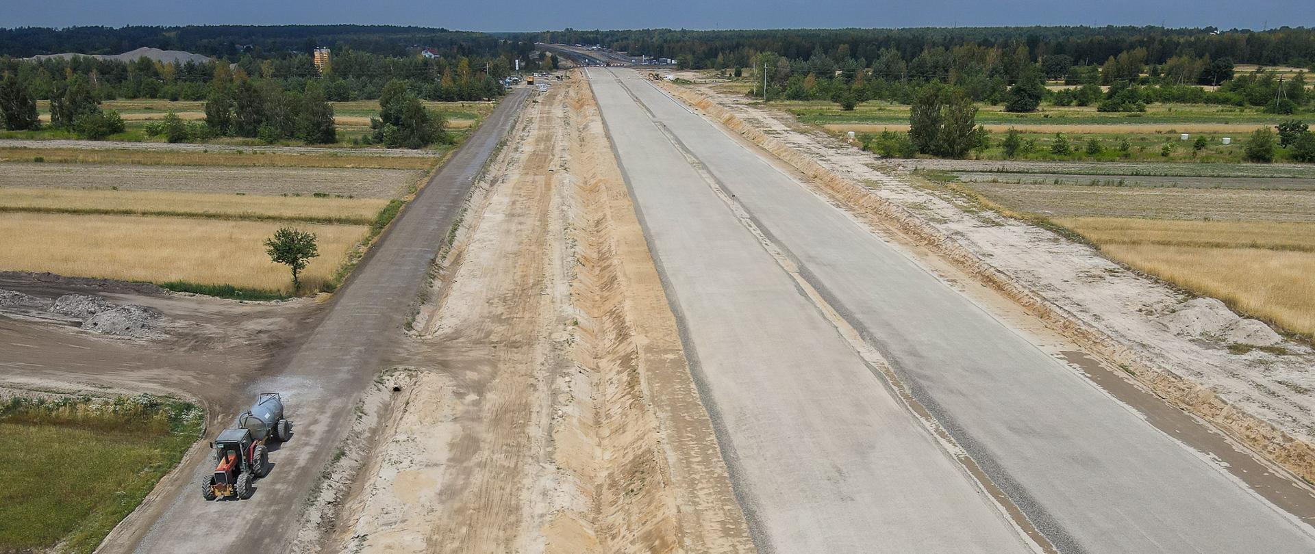 Na zdjęciu widać drogę w budowie oraz jadący traktor, a w oddali widać zieleń.