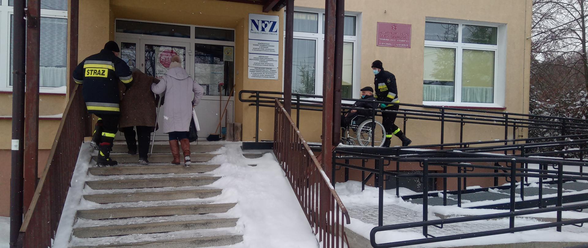 Na zdjęciu widać jak jeden strażak przemieszcza na wózku inwalidzkim jedną osobę do punktu szczepień w msc. Sztabin, drugi strażak wprowadza po schodach (z osobą postronną) drugą osobę.