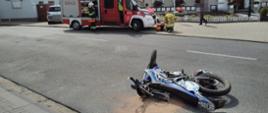 Na zdjęciu widać leżący motocykl oraz w tle wóz strażacki i strażaka 