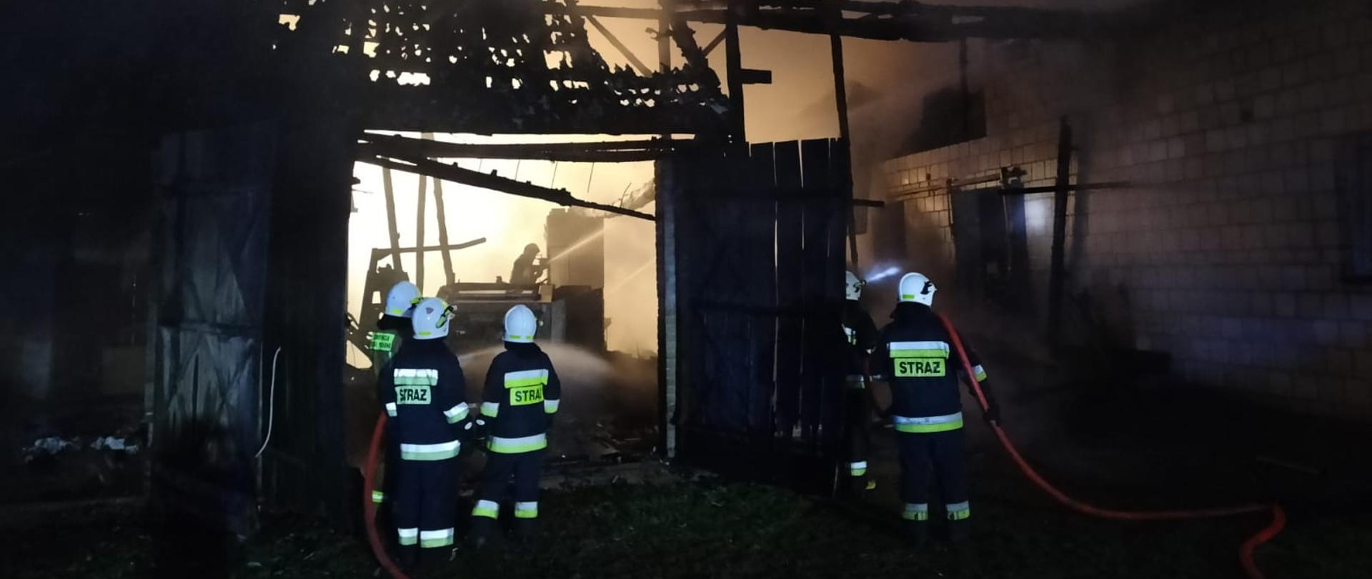 Widok palącego się budynku gospodarskiego z częściowo spalonym dachem. Strażacy stoją z wężami i gaszą pożar.