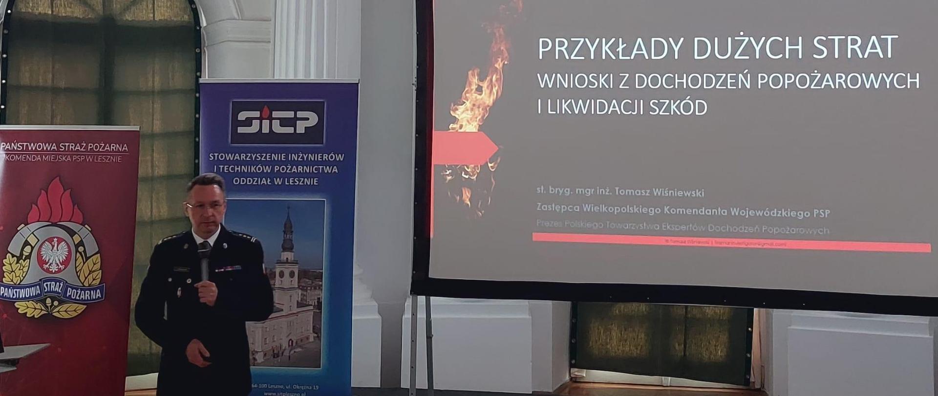 Strażak przemawia do osób siedzących w pomieszczeniu. Obok ekran z prezentacją.
