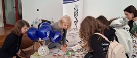 Panie inspektor ubrane w granatowe koszulki z logo PIORiN przedstawiają stoisko promujące inspekcję. Stolik nakryty jest wzorzystym zielono-niebieskim materiałem, na którym leży kratkowany obrus oraz materiały edukacyjne. Dekoracje stoiska stanowią balony, słoiczki oraz foliowe woreczki z nasionami. Obok stoiska znajduje się baner z logo PIORiN. Zainteresowani młodzi ludzie czytają ulotki informacyjne oraz rozpoznają nasiona. 
