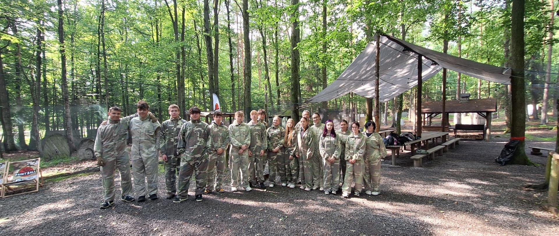 Grupa kilkunastu osób stoi na żwirowym podłożu w lesie, wszyscy w jednakowych, zielonych kombinezonach maskujących do paintballu. Za nimi znajduje się zadaszona wiata z drewnianymi stołami i ławkami. Po lewej stronie widoczny jest pionowy baner z napisem „PAINTBALL”. W tle drzewa o gęstych, zielonych liściach.