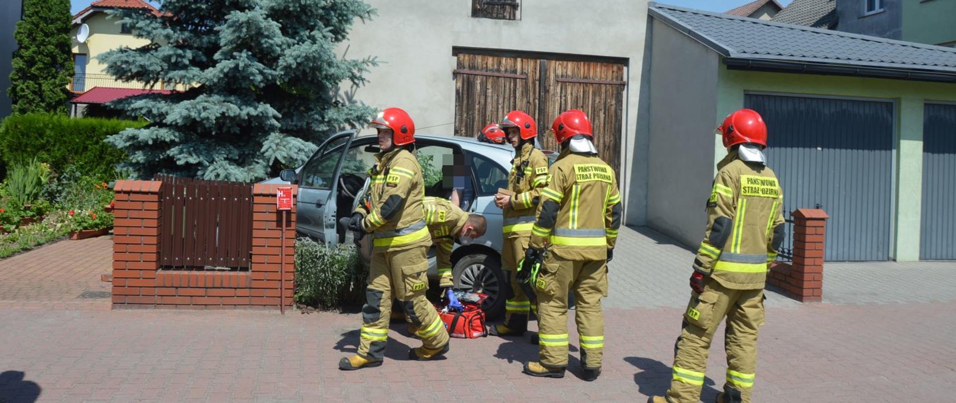 Masłowo, ulica Matejki. Samochód osobowy uderzył w ogrodzenie posesji. Na pierwszym planie strażacy stojący/przemieszczający się wokół auta uczestniczącego w kolizji. W tle zabudowania i drzewa. 