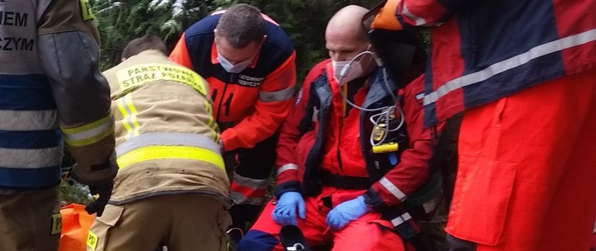 W tle drzewa iglaste. Na środku, na ziemi klęczy strażak w ubraniu specjalnym bez hełmu pochylony nad niewidocznym poszkodowanym. Obok niego po prawej klęczy dwóch ratowników medycznych w uniformach. Obok nich sprzęt medyczny. Skrajnie po lewej stronie stoi strażak w biało niebieskiej kamizelce. Po prawej widoczny stojący ratownik medyczny.