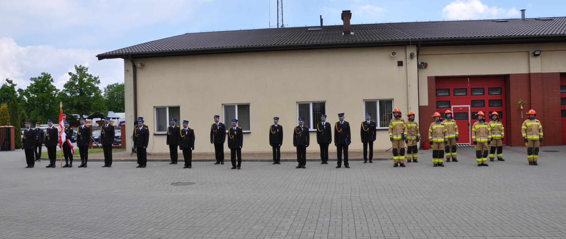 Strażacy stoją na pol-bruku w dwuszeregu na tle żółtego budynku komendy.