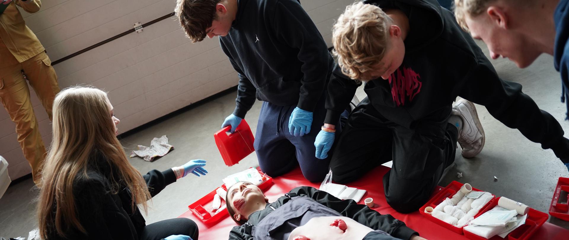 Na zdjęciu widać grupę młodych osób uczestniczących w szkoleniu z pierwszej pomocy. Klęczą wokół leżącej na czerwonej macie osoby, która ma na sobie symulacyjne rany na brzuchu i dłoni. Uczestnicy mają założone rękawiczki ochronne i korzystają z zestawów medycznych z opatrunkami. Jedna osoba instruuje pozostałych, wskazując na poszkodowanego. Scena wygląda na ćwiczenia praktyczne w zamkniętym pomieszczeniu.