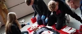 Na zdjęciu widać grupę młodych osób uczestniczących w szkoleniu z pierwszej pomocy. Klęczą wokół leżącej na czerwonej macie osoby, która ma na sobie symulacyjne rany na brzuchu i dłoni. Uczestnicy mają założone rękawiczki ochronne i korzystają z zestawów medycznych z opatrunkami. Jedna osoba instruuje pozostałych, wskazując na poszkodowanego. Scena wygląda na ćwiczenia praktyczne w zamkniętym pomieszczeniu.