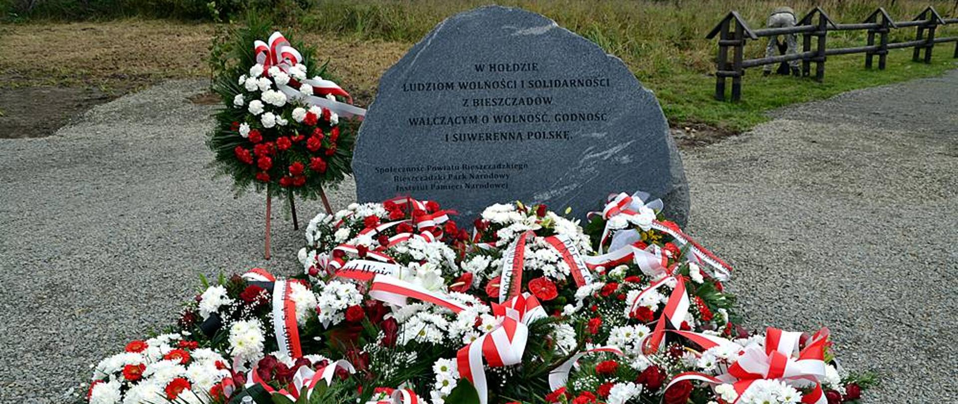 Na zdjęciu widać tablicę upamiętniającą poświęconą ludziom wolności i solidarności z Bieszczad. Pod tablicą złożone są kwiaty oraz wieńce.