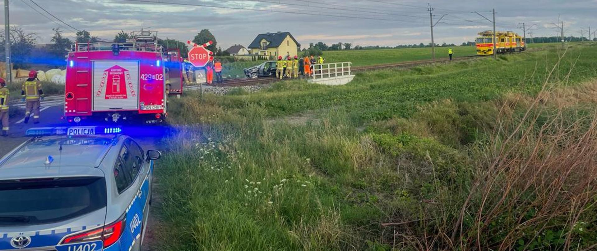 Na zdjęciu widać niestrzeżony przejazd kolejowy na którym doszło do zdarzenia samochodu osobowego z lokomotywą techniczną. W miejscu zdarzenia widać radiowóz policyjny samochód pożarniczy oraz strażaków znajdujących się przy uszkodzonym aucie