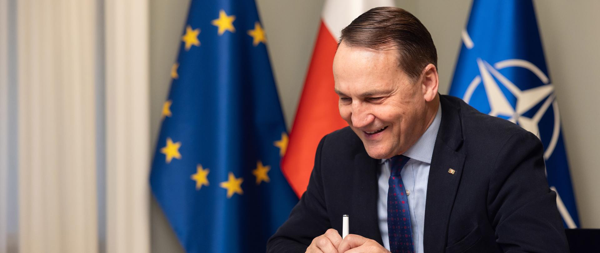 Na zdjęciu jest mężczyzna w garniturze, który siedzi przy stole. Za nim są trzy flagi: Unii Europejskiej, Polski i NATO.