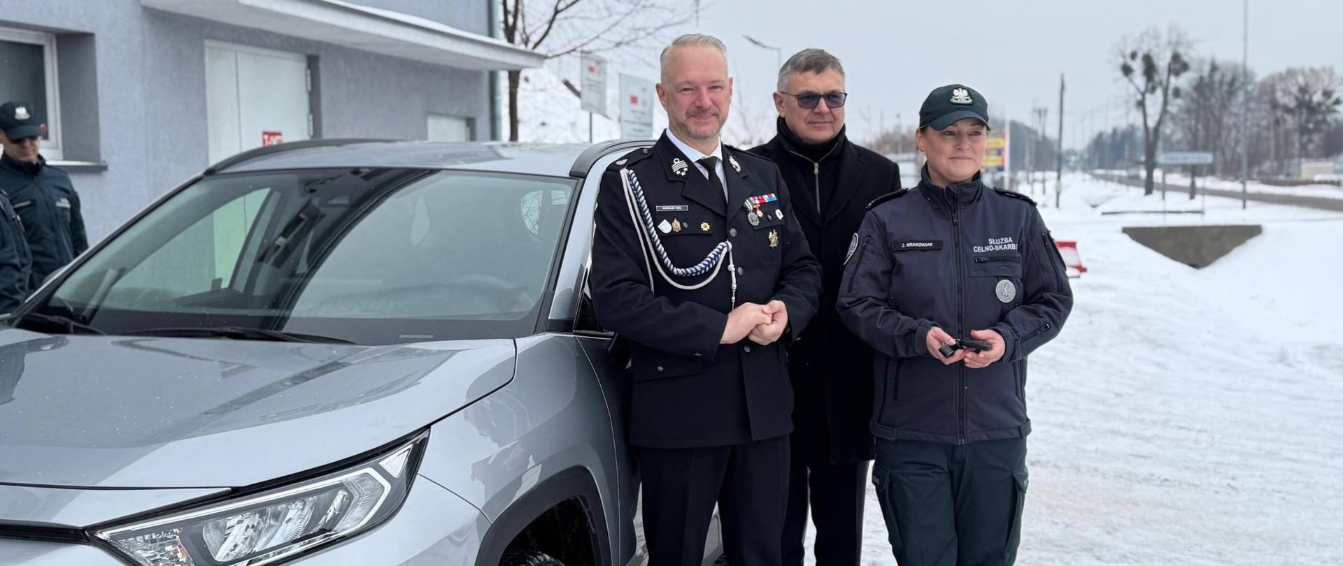 Mężczyzna w galowym mundurze, wojewoda stoi z kobietą w mundurze roboczym oraz mężczyzna w czarnym płaszczu. Stoją przy nowym samochodzie 