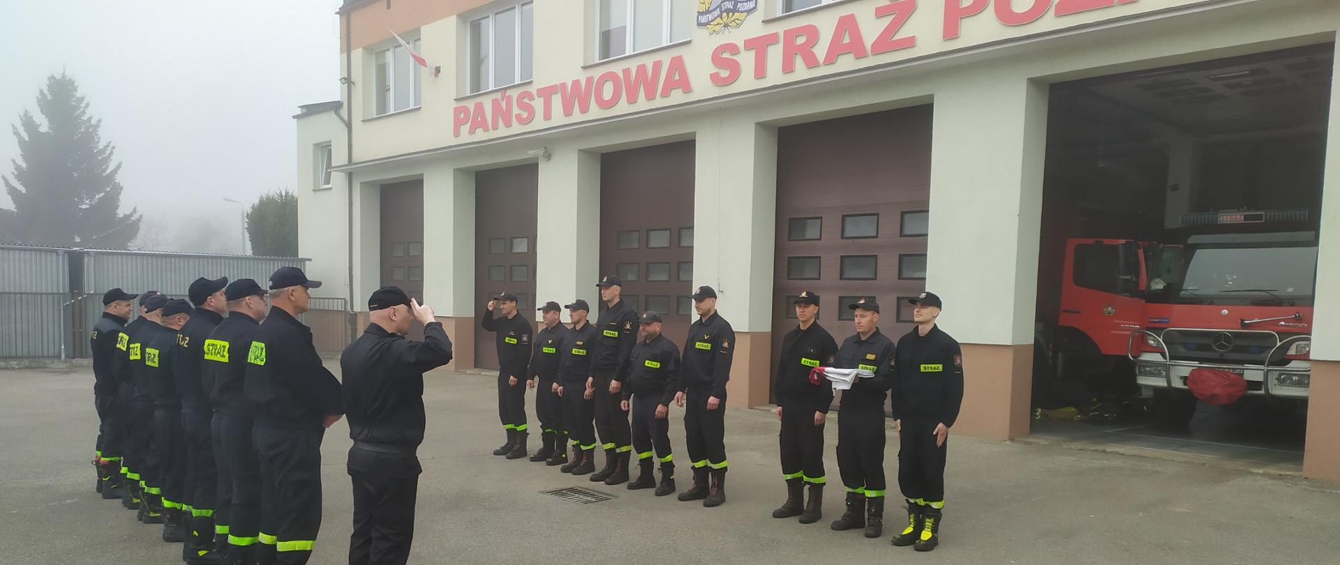 Fotografia przedstawia uroczyste przyjęcie służby z okazji Dnia Flagi Rzeczypospolitej Polskiej. Strażacy z dwóch zmian służbowych stoją na przeciw siebie z prawej strony stoi poczet flagowy składający się z trzech strażaków, z których jeden trzyma złożoną Flagę Państwową w kolorze biało czerwonym.