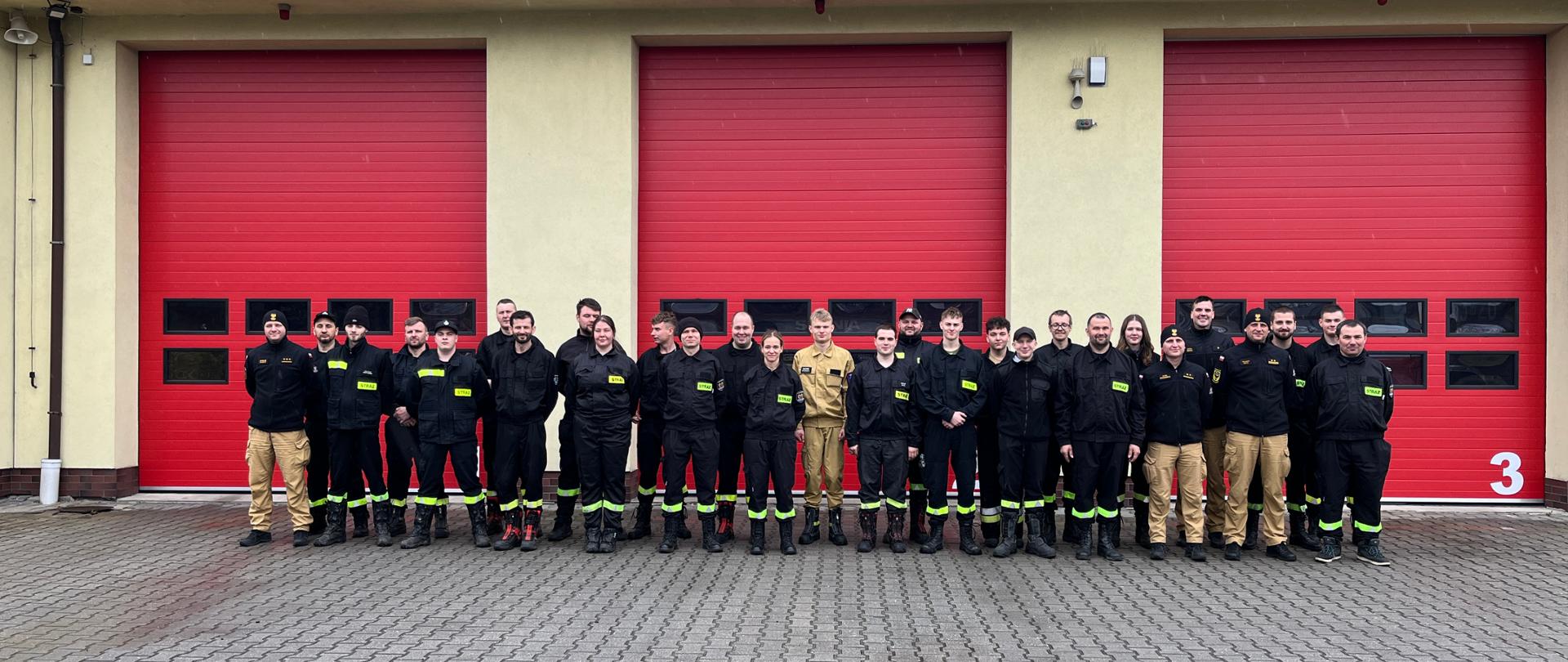 Grupa strażaków i ratowników stoi w dwuszeregu przed budynkiem strażnicy z trzema dużymi czerwonymi bramami garażowymi.