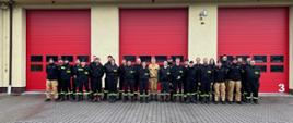 Grupa strażaków i ratowników stoi w dwuszeregu przed budynkiem strażnicy z trzema dużymi czerwonymi bramami garażowymi.