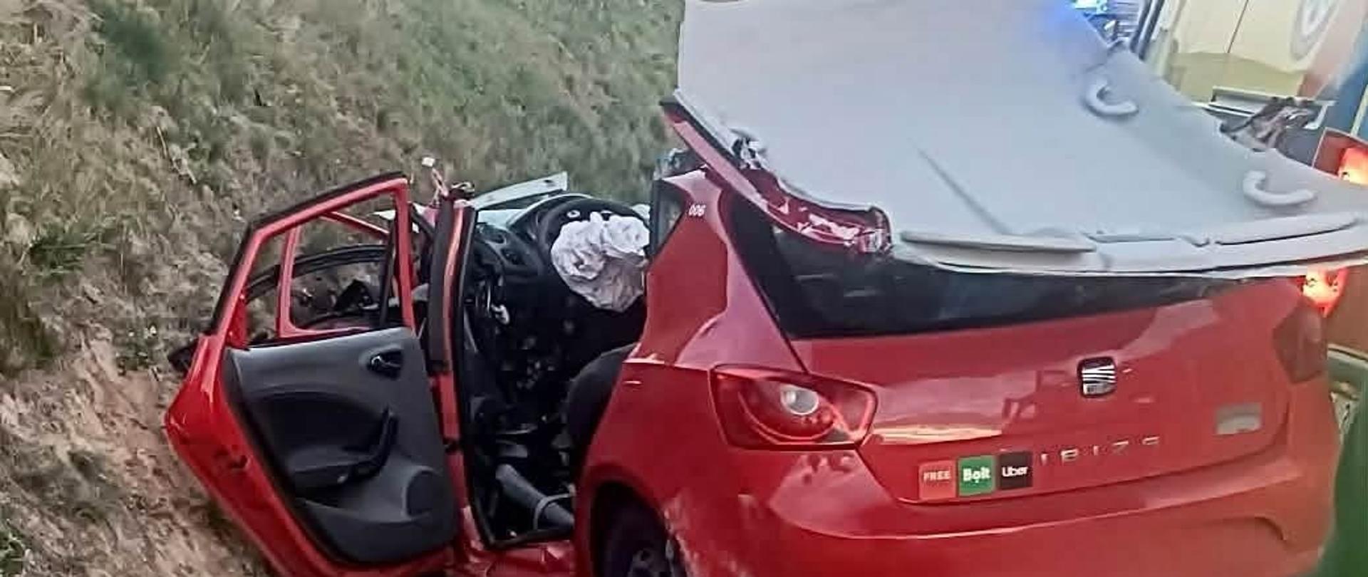 Uszkodzony, czerwony samochód osobowy leży w rowie. Widoczne duże uszkodzenia- zgnieciony przód pojazdu, odcięty i odgięty dach. Boczne drzwi pootwierane, wystrzelona poduszka powietrzna kierowcy. Obok widać kawałek żółtej karetki