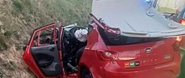 Uszkodzony, czerwony samochód osobowy leży w rowie. Widoczne duże uszkodzenia- zgnieciony przód pojazdu, odcięty i odgięty dach. Boczne drzwi pootwierane, wystrzelona poduszka powietrzna kierowcy. Obok widać kawałek żółtej karetki
