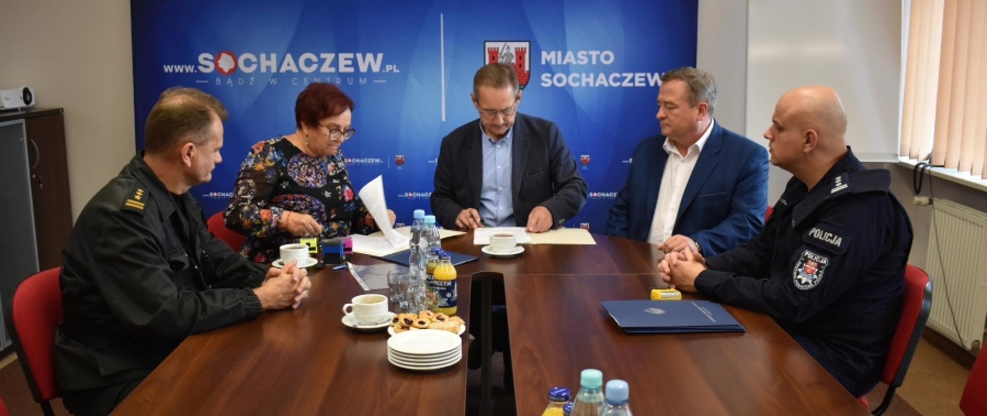 Na zdjęciu widoczne osoby siedzące przy stole, w tym komendanci powiatowi państwowej straży pożarnej i policji. W tle ścianka koloru niebieskiego, z herbem miasta, napisem "Miasto Sochaczew" oraz adresem strony internetowej "www.sochaczew.pl".