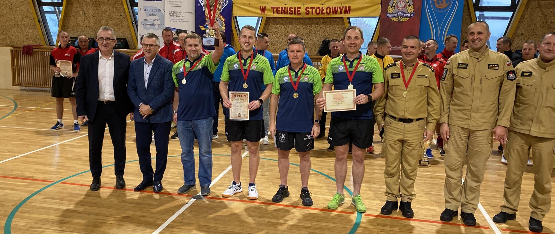 Zdjęcie przedstawia czterech zawodników tenisa stołowego komendy miejskiej PSP w Rzeszowie pozujących z pucharem, medalami oraz dyplomami w asyście gości honorowych przybyłych na zakończenie mistrzostw oraz funkcjonariuszy PSP. W tle widoczne banery PSP, powiatu Strzyżowskiego, klubu strażak, XXIII mistrzostw województwa w tenisie stołowym oraz pozostałe drużyny.