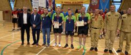 Zdjęcie przedstawia czterech zawodników tenisa stołowego komendy miejskiej PSP w Rzeszowie pozujących z pucharem, medalami oraz dyplomami w asyście gości honorowych przybyłych na zakończenie mistrzostw oraz funkcjonariuszy PSP. W tle widoczne banery PSP, powiatu Strzyżowskiego, klubu strażak, XXIII mistrzostw województwa w tenisie stołowym oraz pozostałe drużyny.