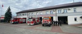 Zdjęcie przedstawia budynek komendy powiatowej psp w Lubaczowie. Przed budynkiem widoczne są pojazdy ratowniczo-gaśnicze oraz strażacy PSP którzy stoją w szeregu przed garażami. 