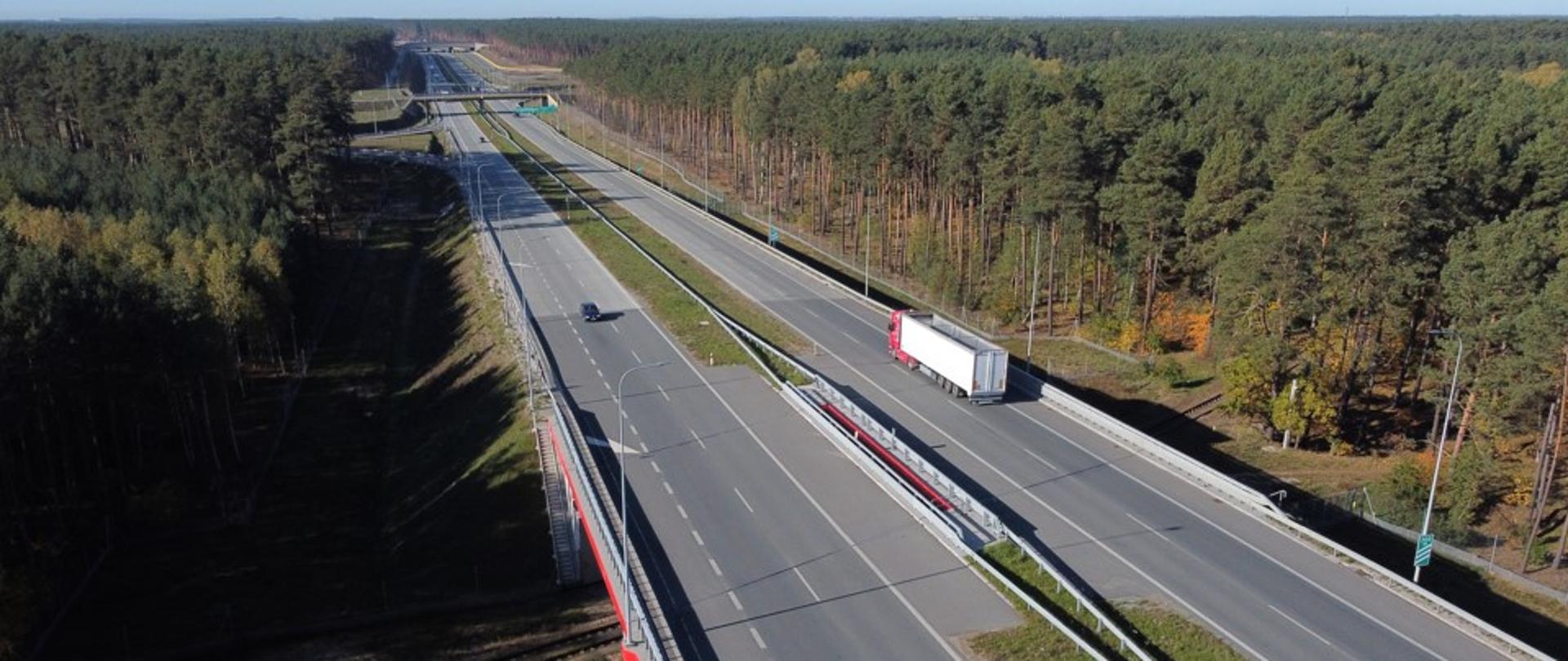Dwujezdniowa droga biegnąca przez las. Widoczny wiadukt nad linią kolejową. Słoneczna pogoda