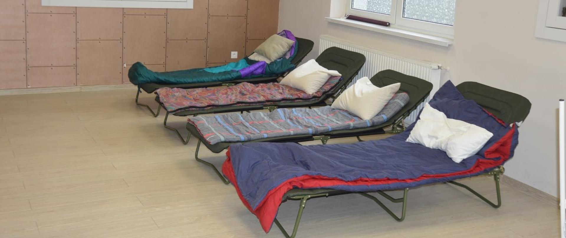 Miejsca noclegowe w siedzibie OSP. W salce szkoleniowych znajdują się 4 łózka polowe. Na nich leżą śpiwory i poduszki. Na ścianie w gablocie jest zawieszony sztandar strażacki.