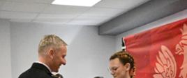 Strażak w mundurze wyjściowym gratuluje dziewczynie i wręcza dyplom. W prawej części zdjęcia czerwony baner PSP. W tle przed białą ścianą stoją inni uczestnicy turnieju. 