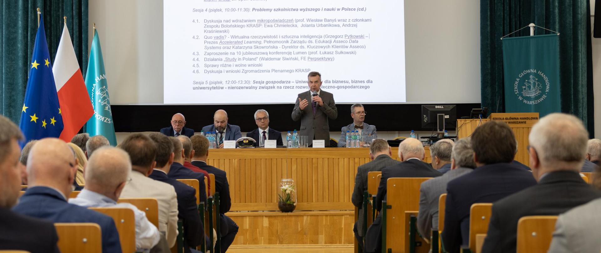 Widok od tyłu na salę, na ustawionych w rzędy krzesłach siedzą ludzie, przed nimi na podwyższeniu za drewnianym stołem stoi minister Wieczorek i mówi do mikrofonu, obok niego siedzi kilku mężczyzn w garniturach.