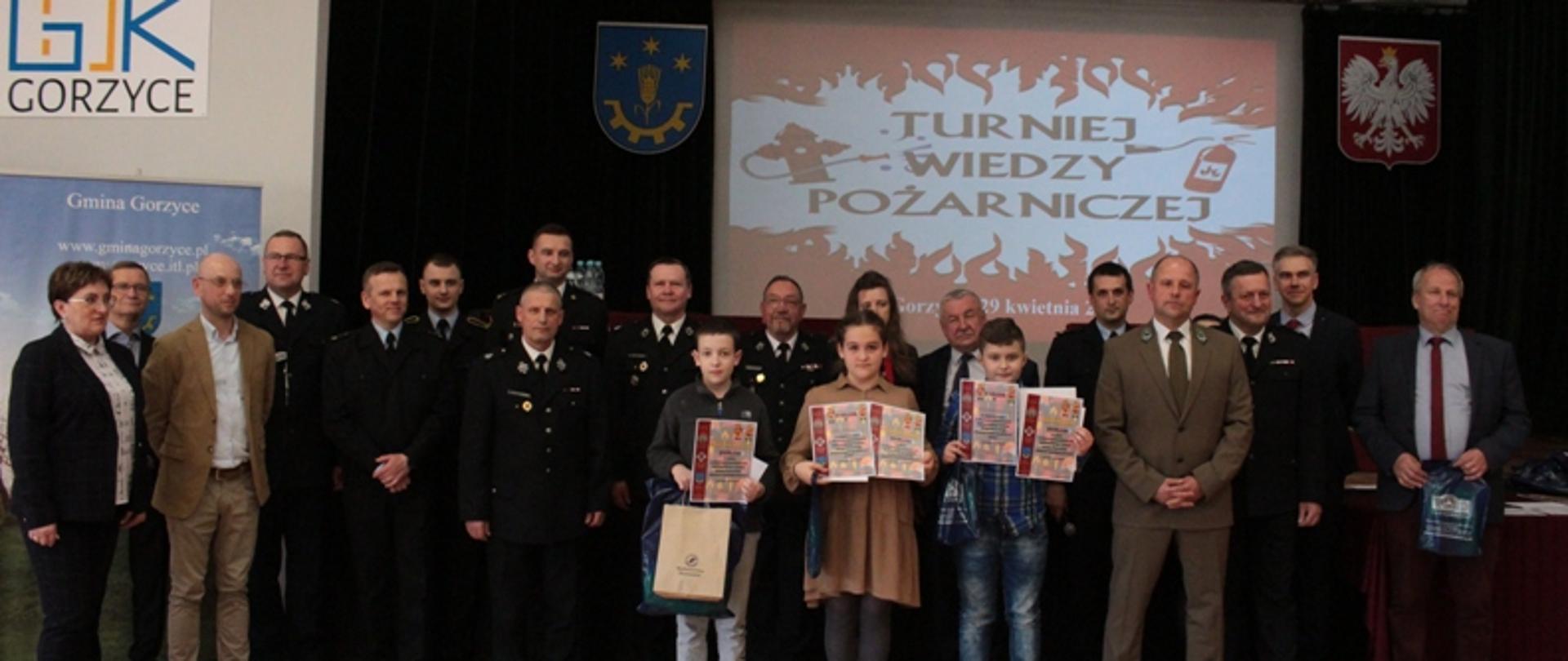 Grupa osób w dużym pomieszczeniu pozuje do fotografii. W pierwszym rzędzie stoją dzieci, które są uczestnikami turnieju wiedzy pożarniczej, za nimi stoją obserwatorzy turnieju.