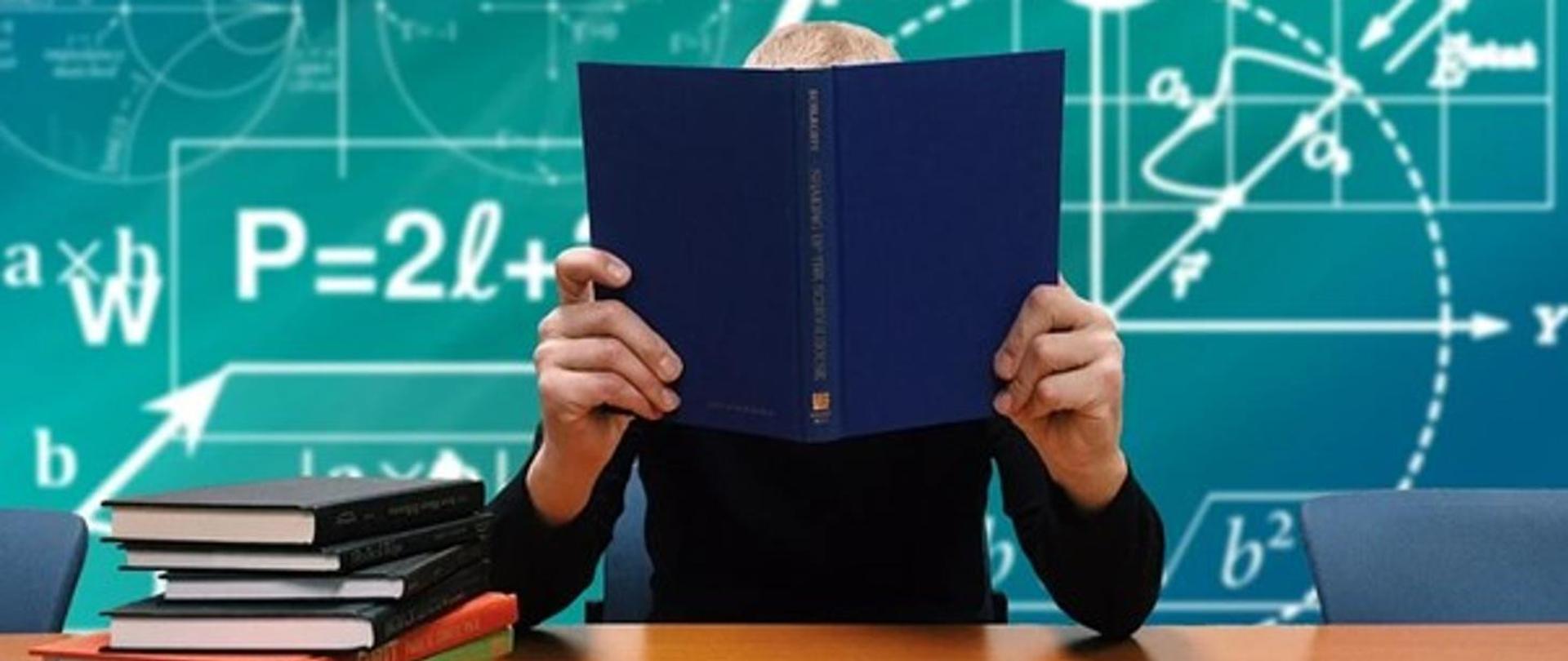 Nauczyciel siedzi pzry biurku w szkolnej klasie i trzyma otwarty podręcznik na wysokści twarzy.na tle tablicy szkonej