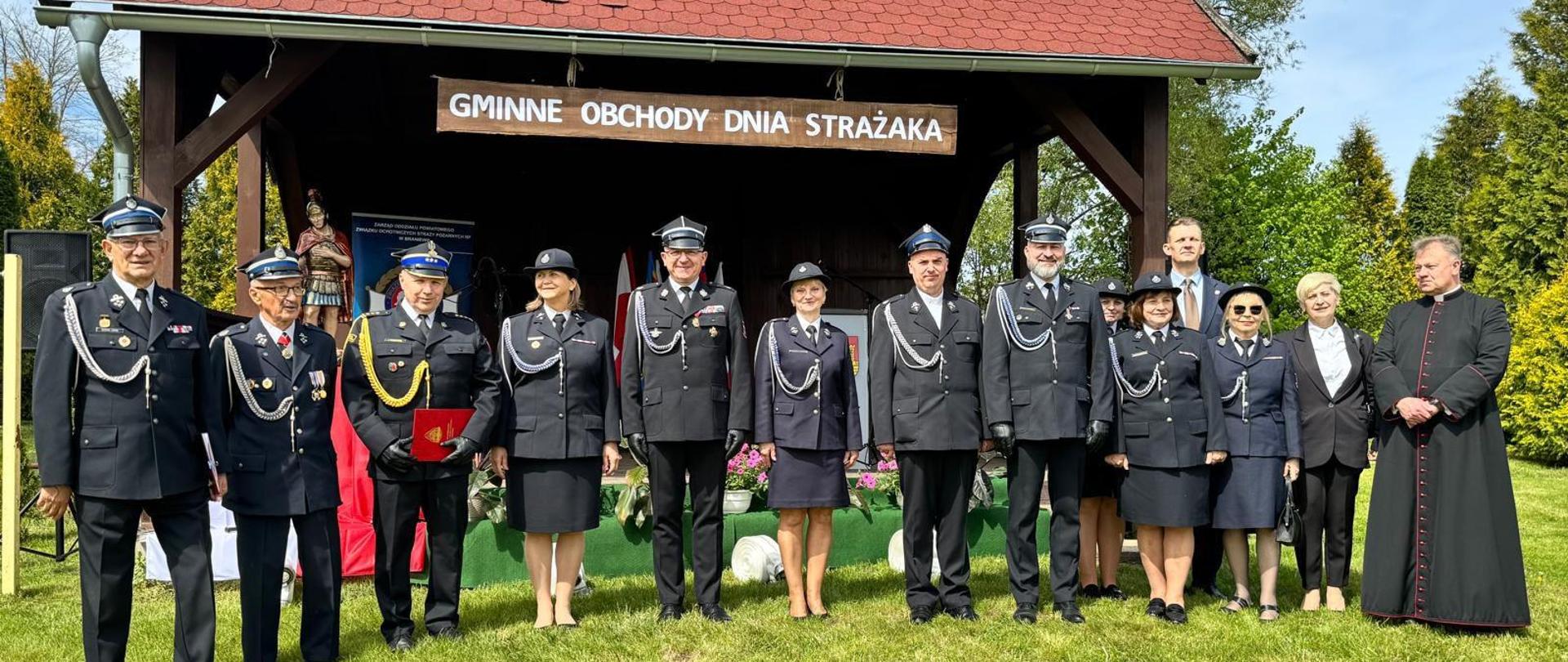 Zielona polana. Na zdjęciu grupa strażaków w mundurach galowych. Z prawej strony stoi duchowny. W tle wiata ze sceną.