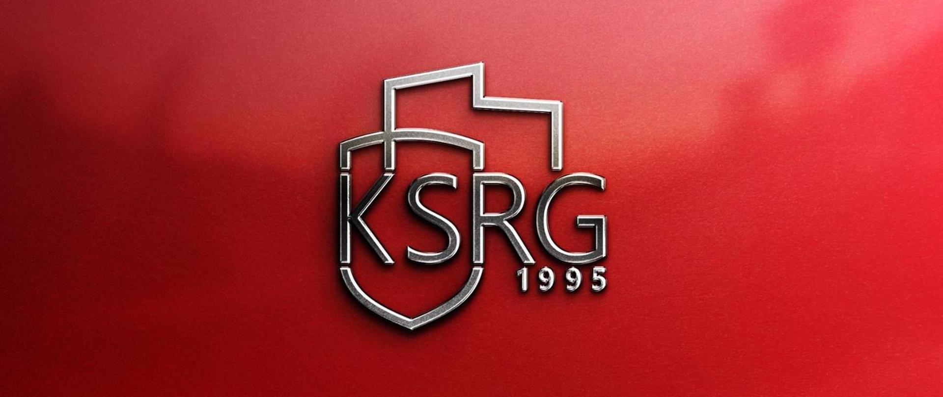 Obraz przedstawia trójwymiarowe, metaliczne logo "KSRG" na czerwonym, gradientowym tle. Litery "KSRG" są stylizowane i wpisane w tarczę herbową, nad którą znajduje się stylizowany zarys dachu. Pod literami "KSRG", po prawej stronie, umieszczono cyfry "1995". Logo ma srebrzysty, błyszczący wygląd, który sugeruje efekt metalu. Czerwone tło jest jaśniejsze po prawej stronie i ciemniejsze po lewej, z delikatnym efektem winiety.
