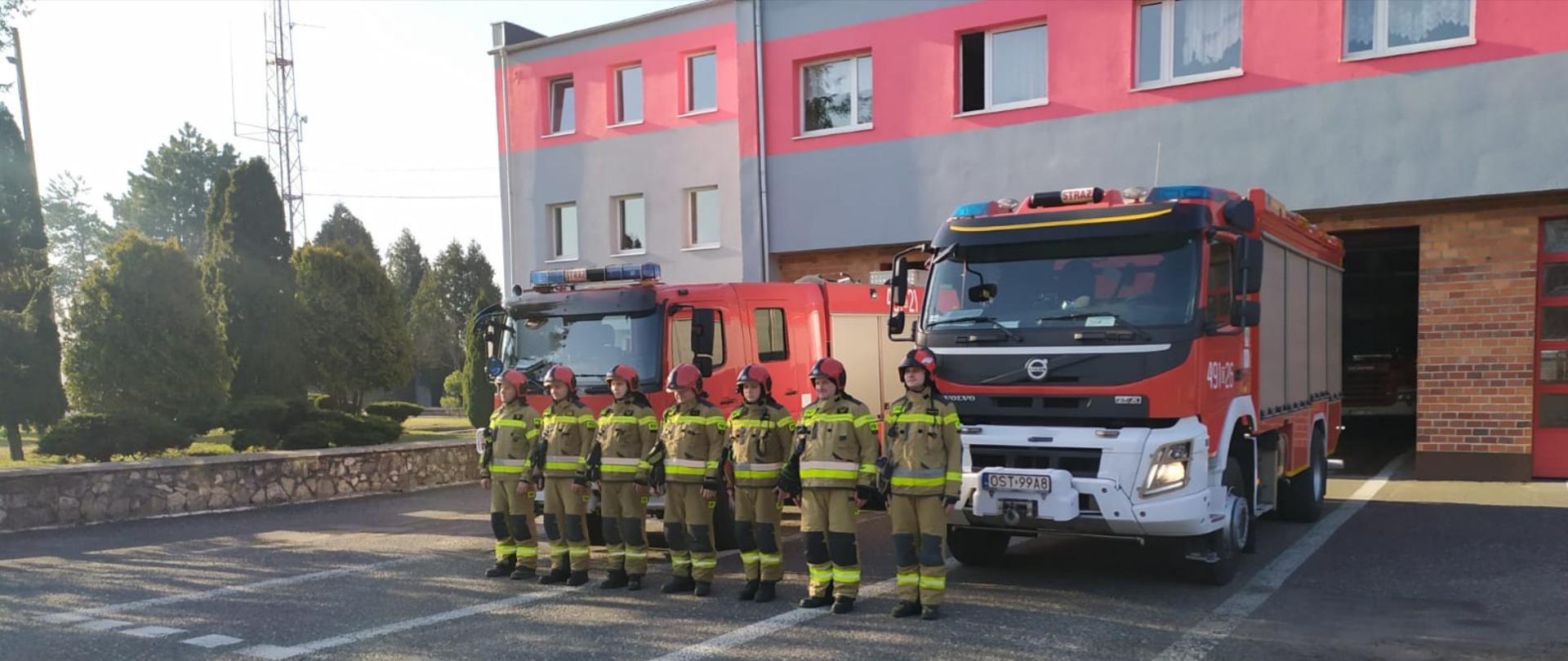 Na zdjęciu znajdują się strażacy podczas zbiórki na tle pojazdów i budynku