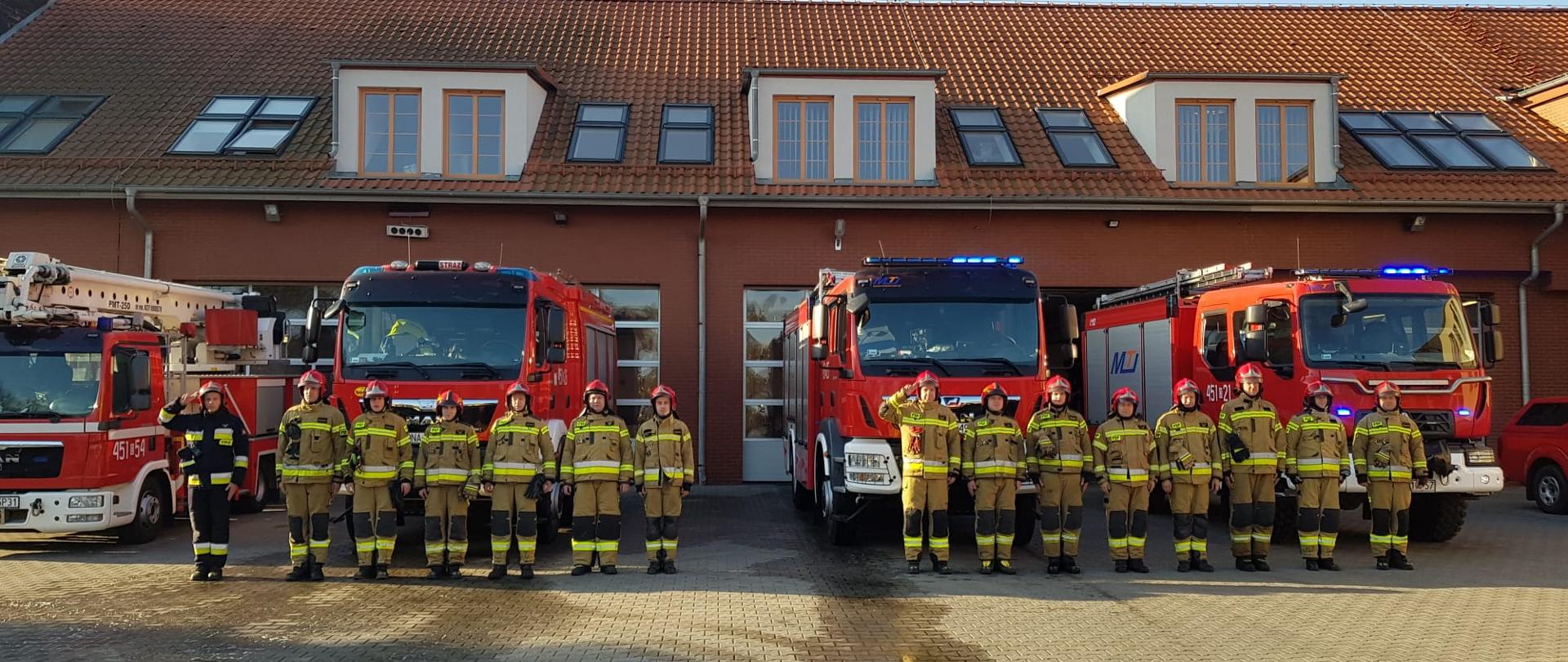 Strażacy PSP w umundurowaniu specjalnym stoją przed pojazdami pożarniczymi koloru czerwonego, które stoją przed remizami JRG PSP. Strażacy oddają hołd poległym strażakom w Ukrainie.