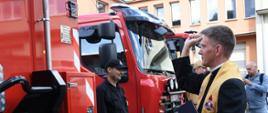 Kapelan święci kontener sanitarny obok stoi strażak w mundurze koloru czarnego obok stoją dwa samochody gaśnicze.