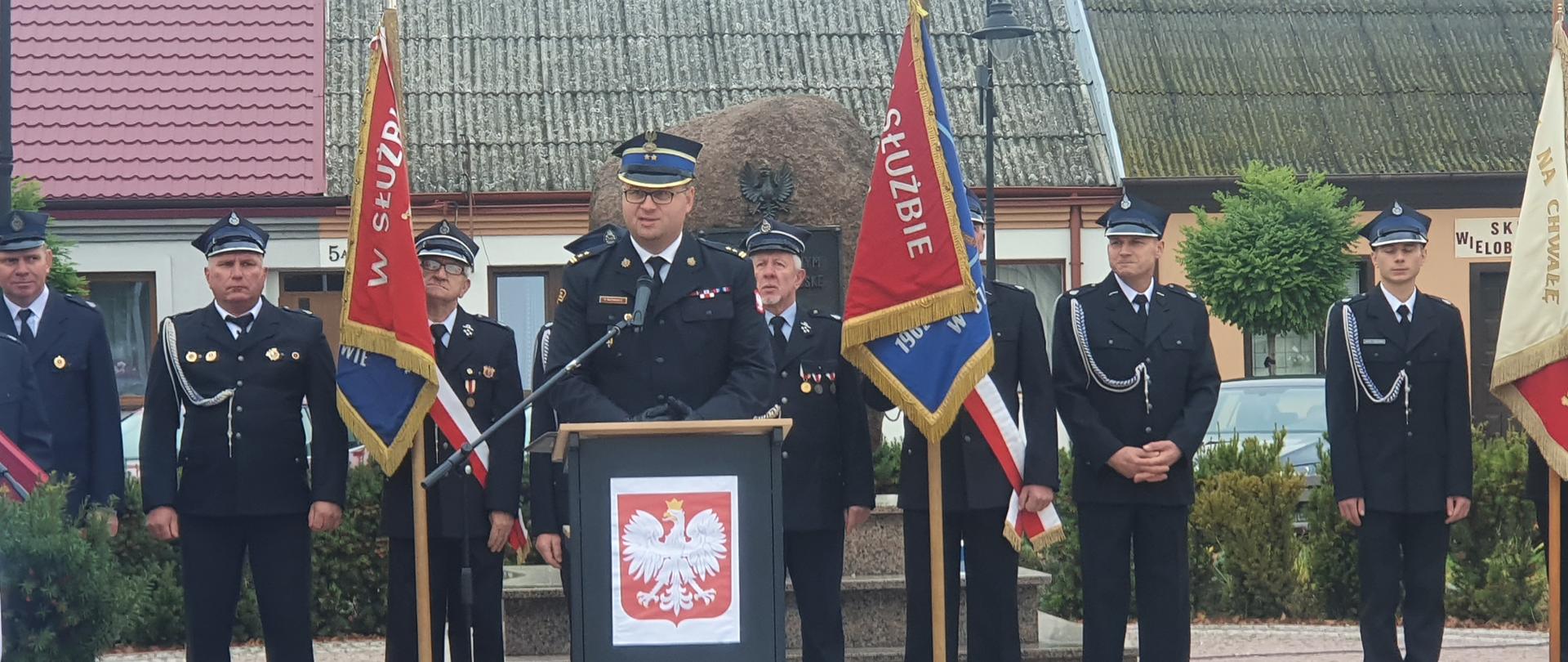 strażak w rogatywce stoi przy mównicy, za nim stoi grupa strażakó w mundurach i w rogatywkach