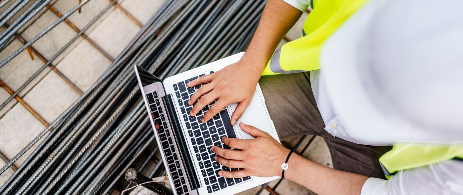 Osoba w kasku i kamizelce odblaskowej pracuje na laptopie na placu budowy; w tle zbrojenie i betonowe elementy.