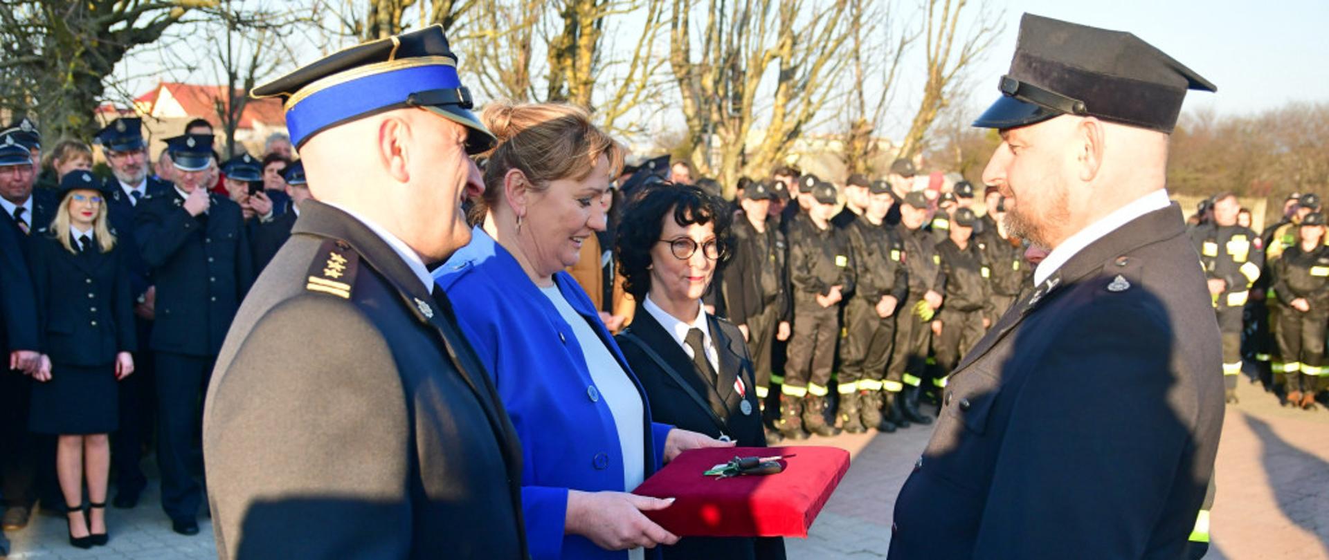 Na zdjęciu na pierwszym planie widać czwórkę ludzi. P.o. Kujawsko-Pomorski Komendant Wojewódzki PSP w Toruniu, Wójt Gminy Lubiewo oraz Przewodnicząca Rady Gminy Lubiewo stoją naprzeciwko Prezesa OSP Lubiewo. P.o. Kujawsko-Pomorski Komendant Wojewódzki PSP w Toruniu trzyma w ręku czerwoną teczkę oraz pluszowego misia. Pani Wójt Gminy Lubiewo wręcza na czerwonej poduszce klucze od pojazdy prezesowi OSP Lubiewo. Przewodnicząca Rady Gminy Lubiewo 