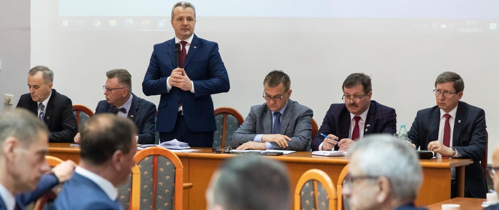 Jeden mężczyzna stojący z mikrofonem w ręku i wypowiadający się, wielu mężczyzn siedzących przy stole