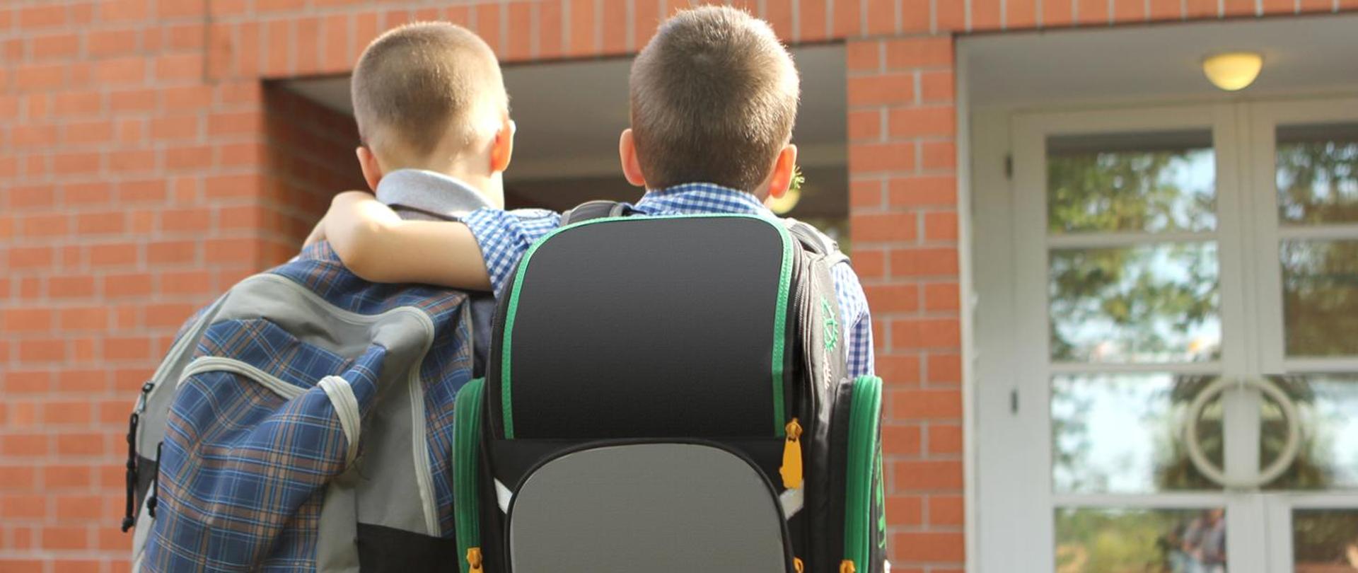 dwóch chłopców w wieku szkolnym odwróconych tyłem do obiektywu z plecakami na plecach. Tło stanowi wejście do budynku najprawdopodobniej szkoły.
