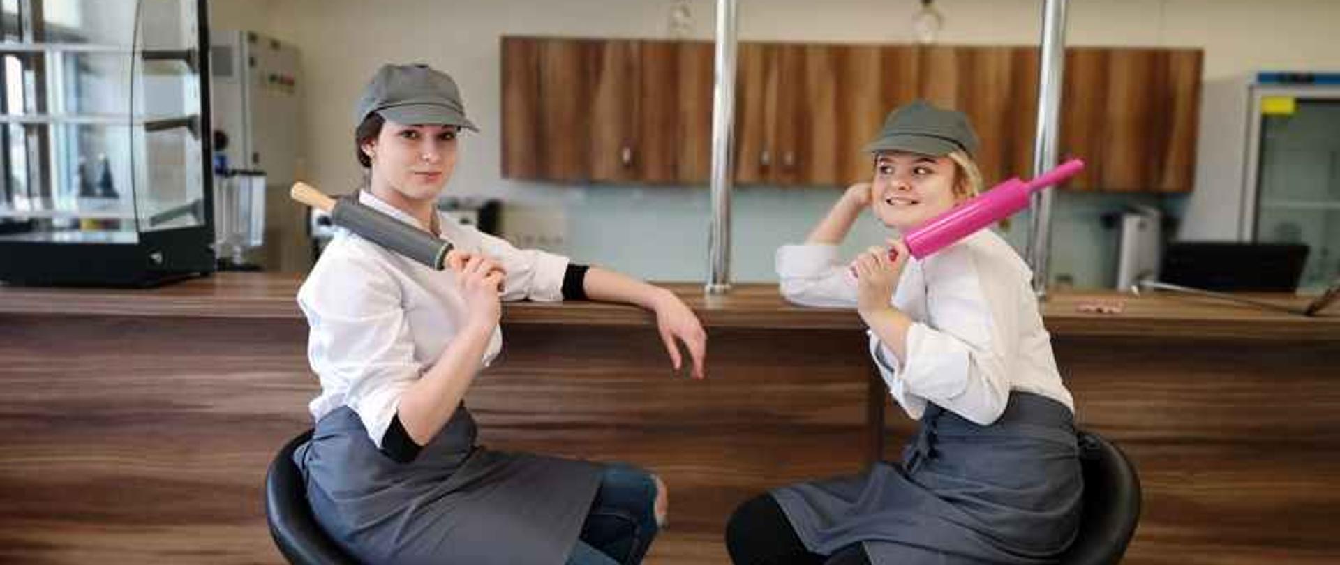 Przy barze siedzą dwie młode dziewczyny. Każda z nich trzyma wałek do ciasta.