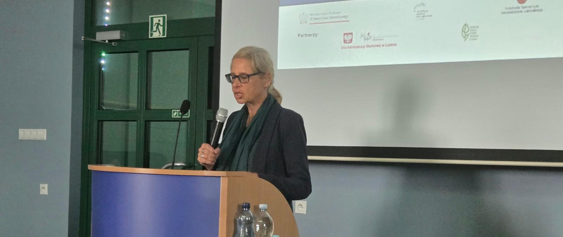 Na zdjęciu widoczna jest kobieta przemawiająca przy mównicy podczas konferencji. W tle znajduje się ekran projekcyjny z prezentacją, a obok ustawiono stół z komputerem, materiałami oraz butelkami wody. Na froncie mównicy widoczny jest napis „Muzeum Badań Polarnych w Puławach”.
