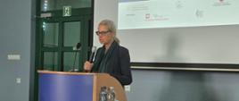 Na zdjęciu widoczna jest kobieta przemawiająca przy mównicy podczas konferencji. W tle znajduje się ekran projekcyjny z prezentacją, a obok ustawiono stół z komputerem, materiałami oraz butelkami wody. Na froncie mównicy widoczny jest napis „Muzeum Badań Polarnych w Puławach”.